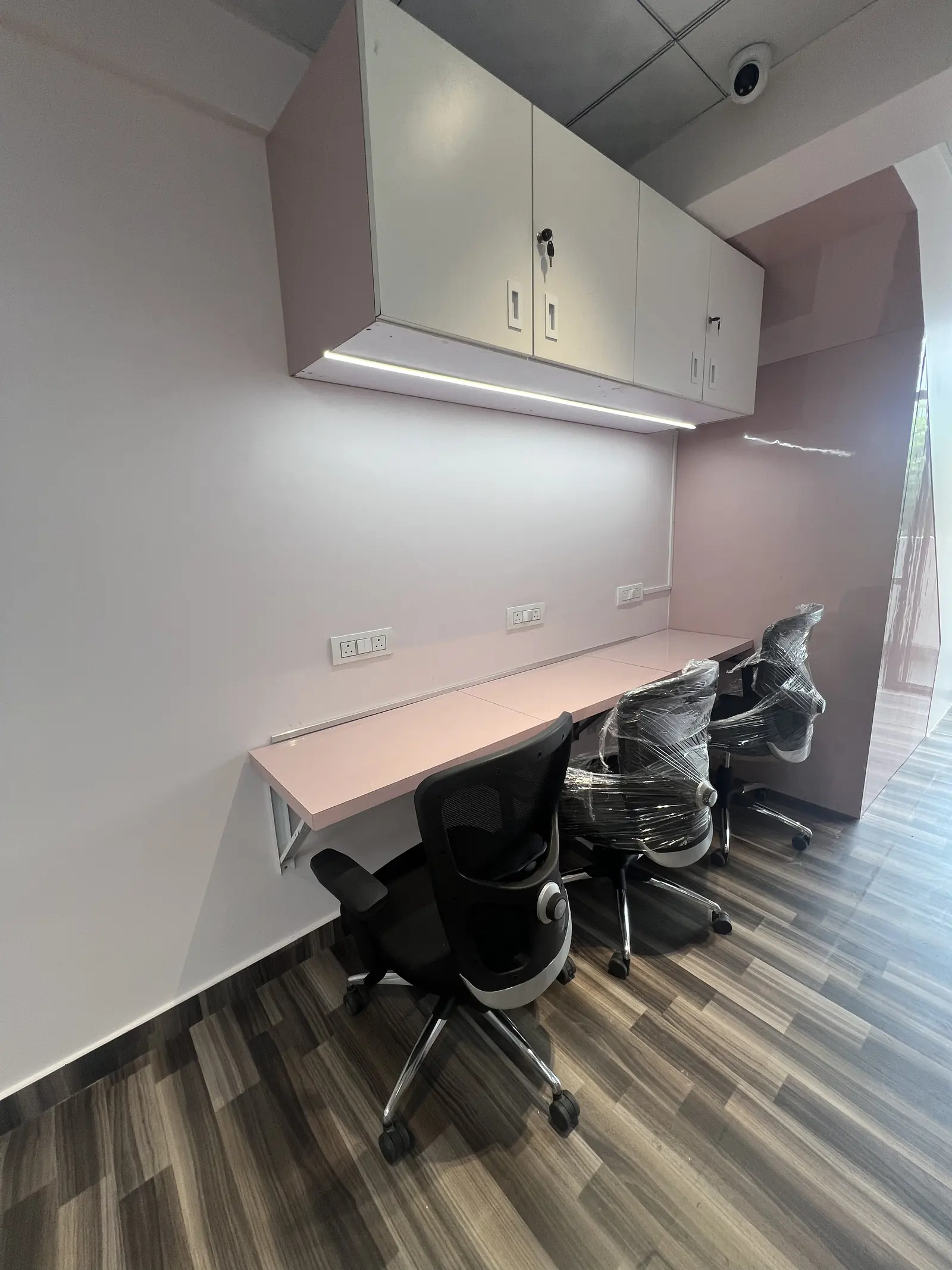 A row of blush-pink desks with black mesh chairs and lockable overhead cabinets